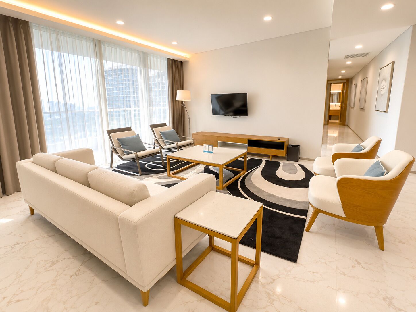 A modern hotel room with a sofa, two armchairs, a coffee table, a TV, and a large window with curtains.