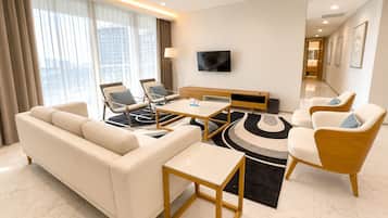 A modern hotel room with a sofa, two armchairs, a coffee table, a TV, and a large window with curtains.
