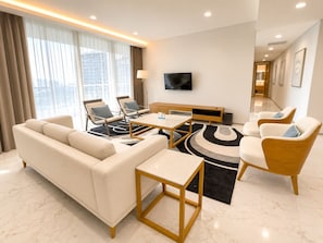 A modern hotel room with a sofa, two armchairs, a coffee table, a TV, and a large window with curtains.