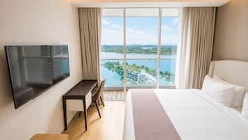 A hotel room with a bed, a desk, a TV, and a large window with a view of a lake and buildings.