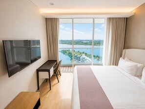 Ein Hotelzimmer mit einem Bett, einem Schreibtisch, einem Fernseher und einem großen Fenster mit Blick auf einen See und Gebäude.