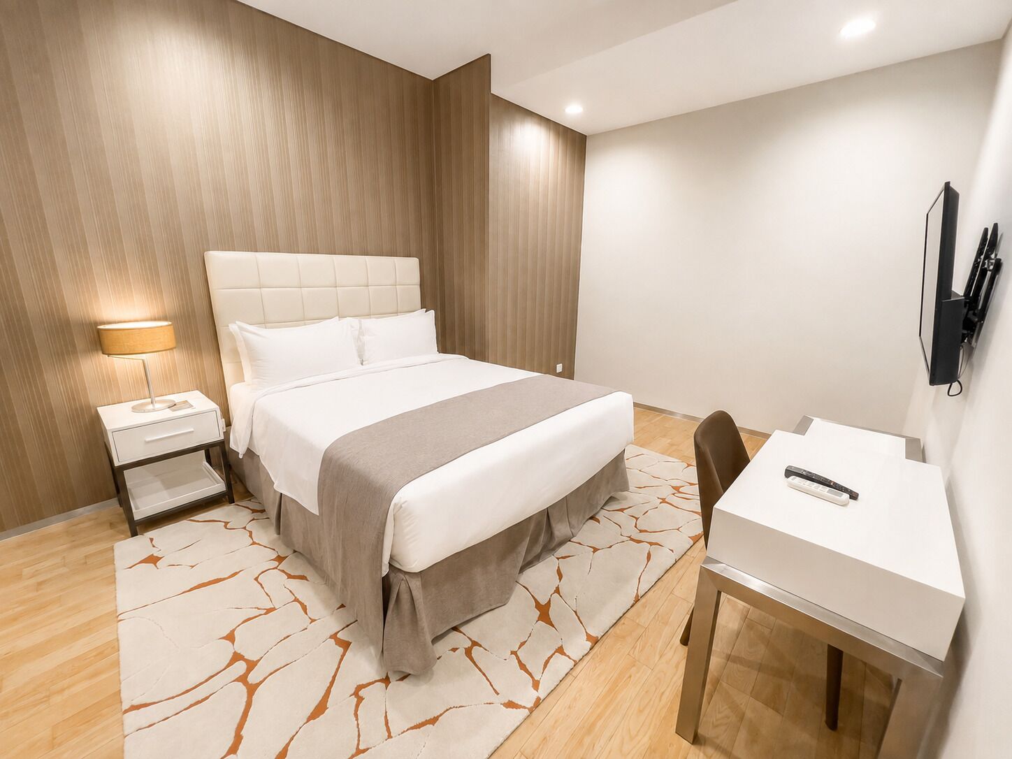 A hotel room with a bed, bedside table, desk, chair, and a TV mounted on the wall.