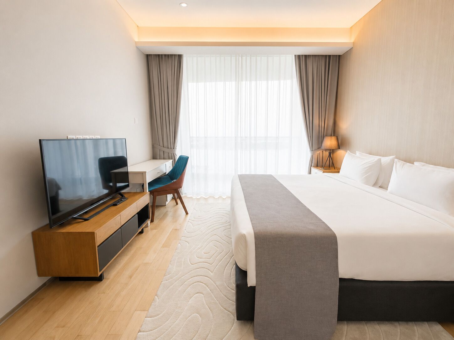 A hotel room with a bed, a desk with a television, a chair, a lamp, and a window with curtains.
