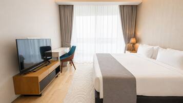 A hotel room with a bed, a desk with a television, a chair, a lamp, and a window with curtains.
