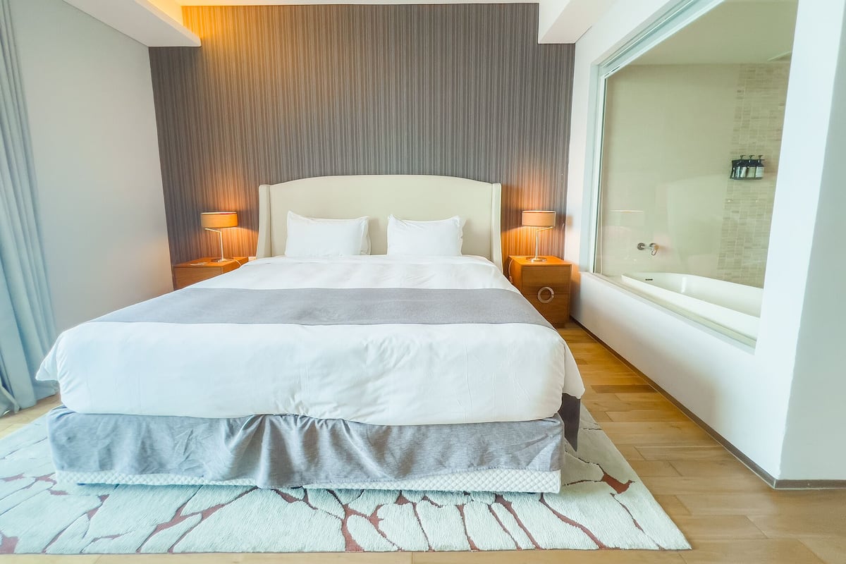 A modern hotel room with a large bed, two bedside tables with lamps, a glass shower enclosure, and a view of the city.