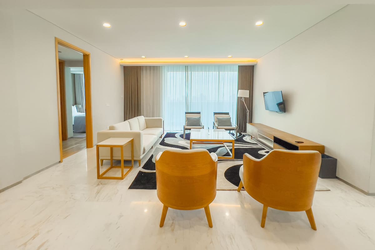 A modern hotel room with a sofa, two armchairs, a coffee table, a TV, and a large window with curtains.