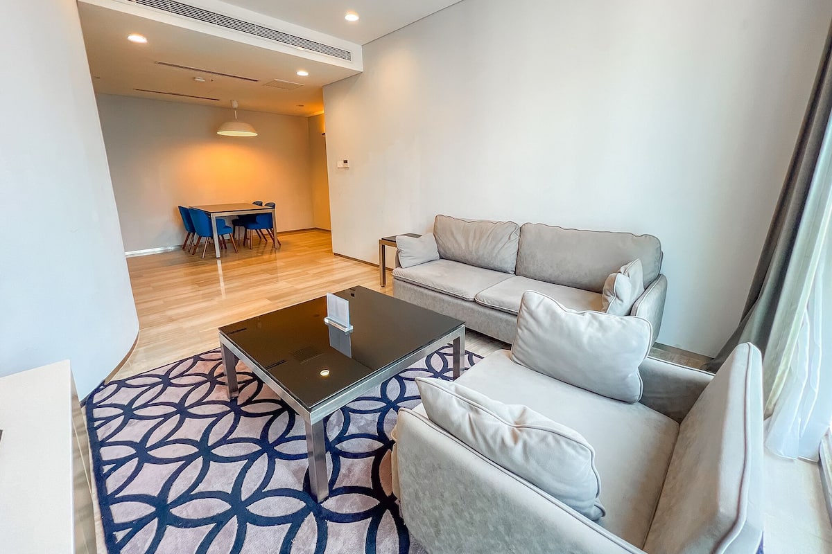 A modern living room with a sofa, a glass coffee table, and a patterned rug. There is a dining area with a table and chairs in the background.