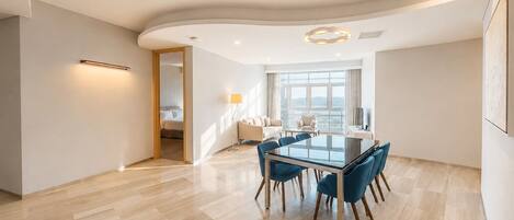 A modern hotel room with a dining area featuring a glass table and blue chairs, a large window with a view, and a curved ceiling design.