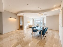 Et moderne hotelværelse med et spiseområde, der inkluderer et glasbord og blå stole, et stort vindue med udsigt og et buet loftdesign.