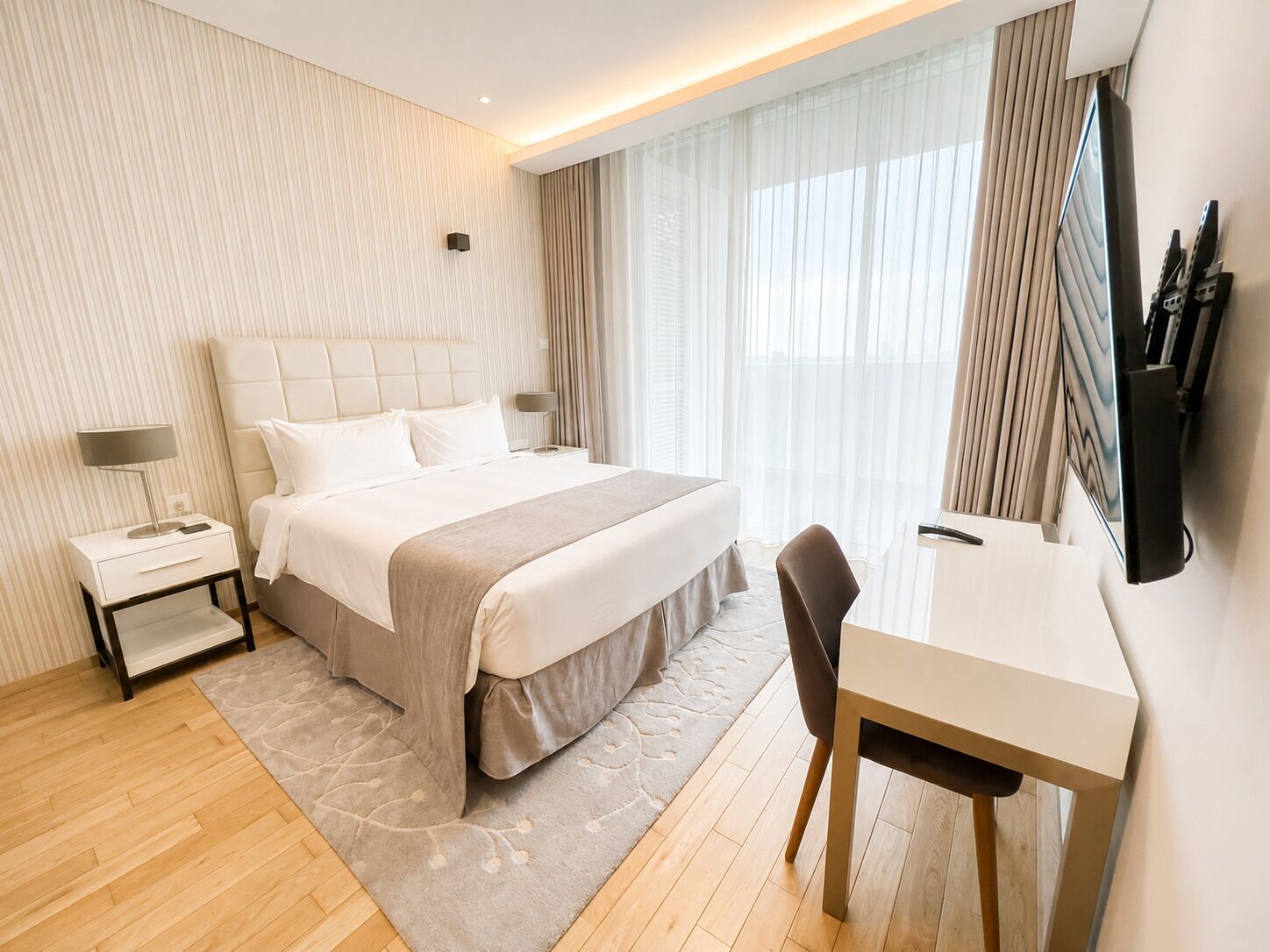 A hotel room with a bed, bedside table, lamp, desk, chair, and a TV mounted on the wall.