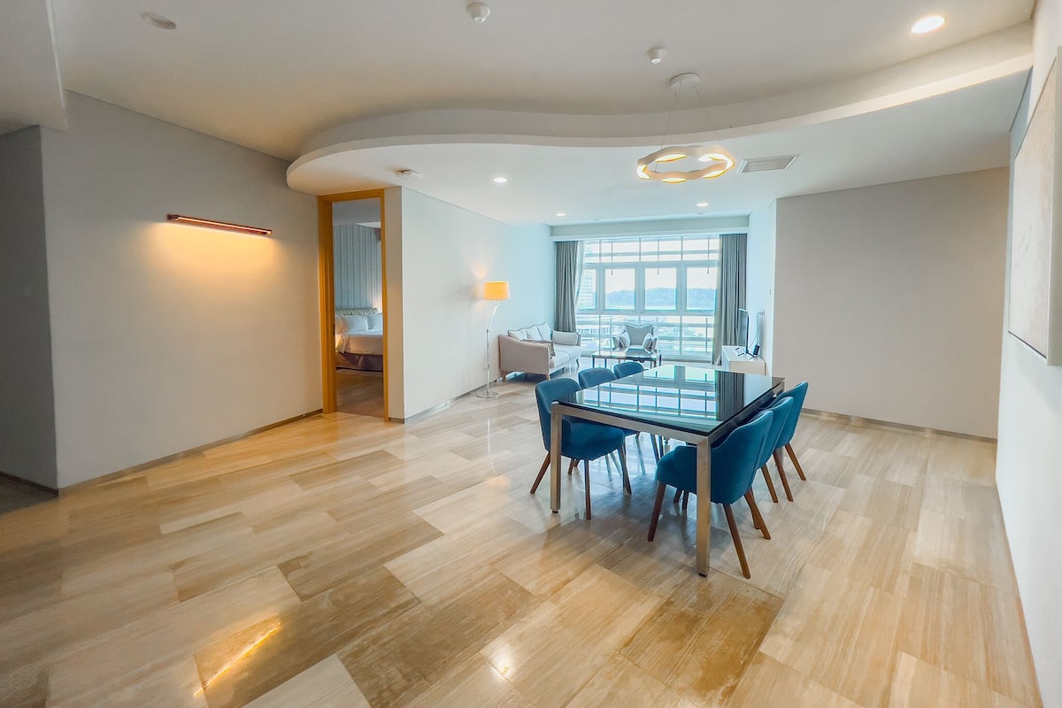 A modern hotel room with a dining area featuring a glass table and blue chairs, a large window with a view, and a curved ceiling with recessed lighting.