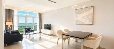 A modern hotel room with a dining area, a sofa, a TV, and a large window offering a scenic view.
