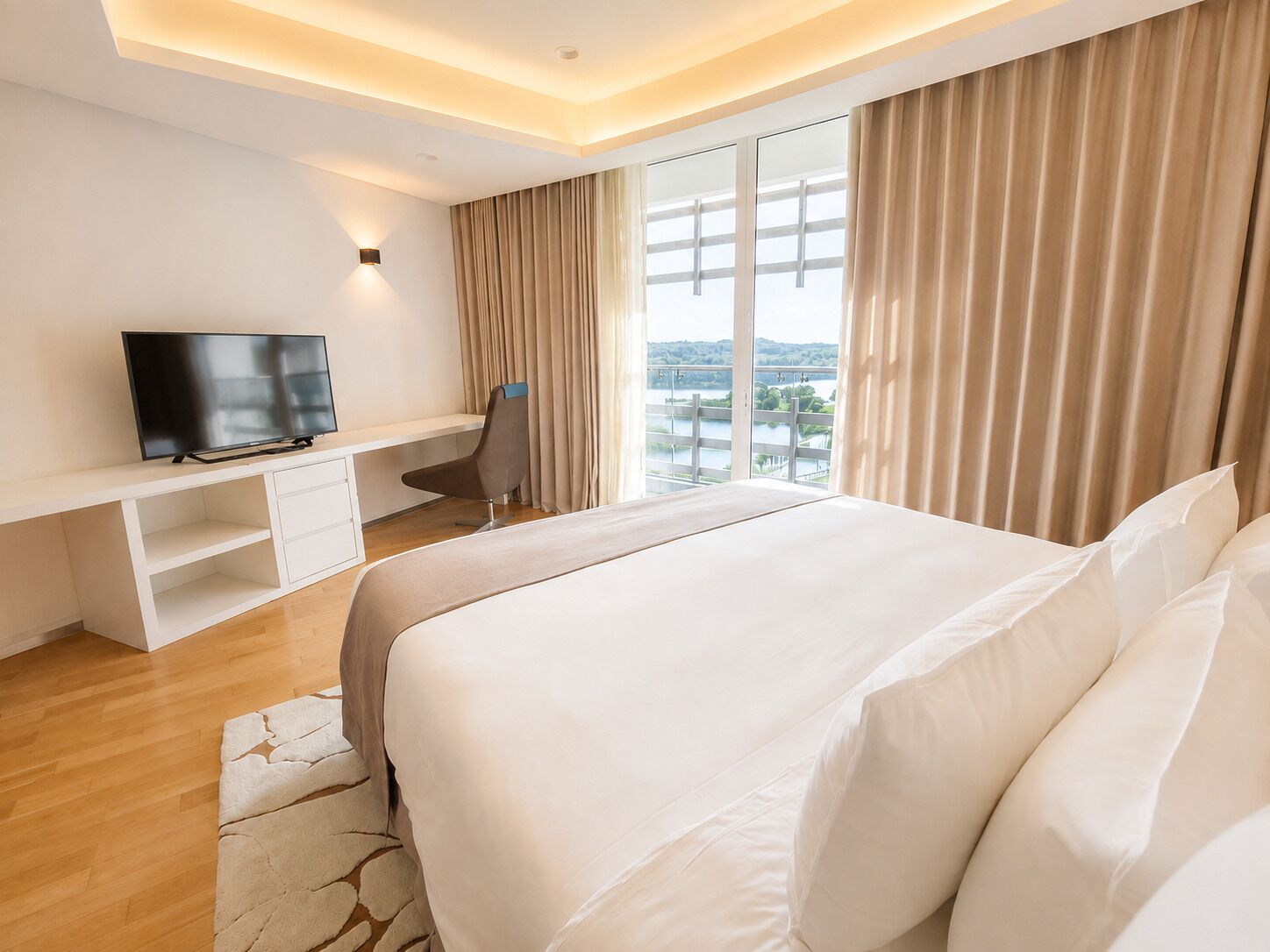 A hotel room with a large bed, a television on a white console, a desk, and a view of a body of water through large windows.