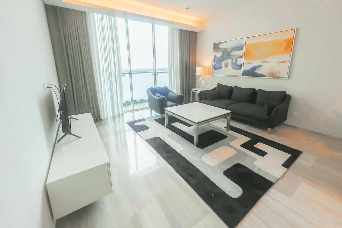 A modern living room with a white coffee table, a black sofa, and a large window with curtains. There are abstract paintings on the wall and a lamp on a side table.