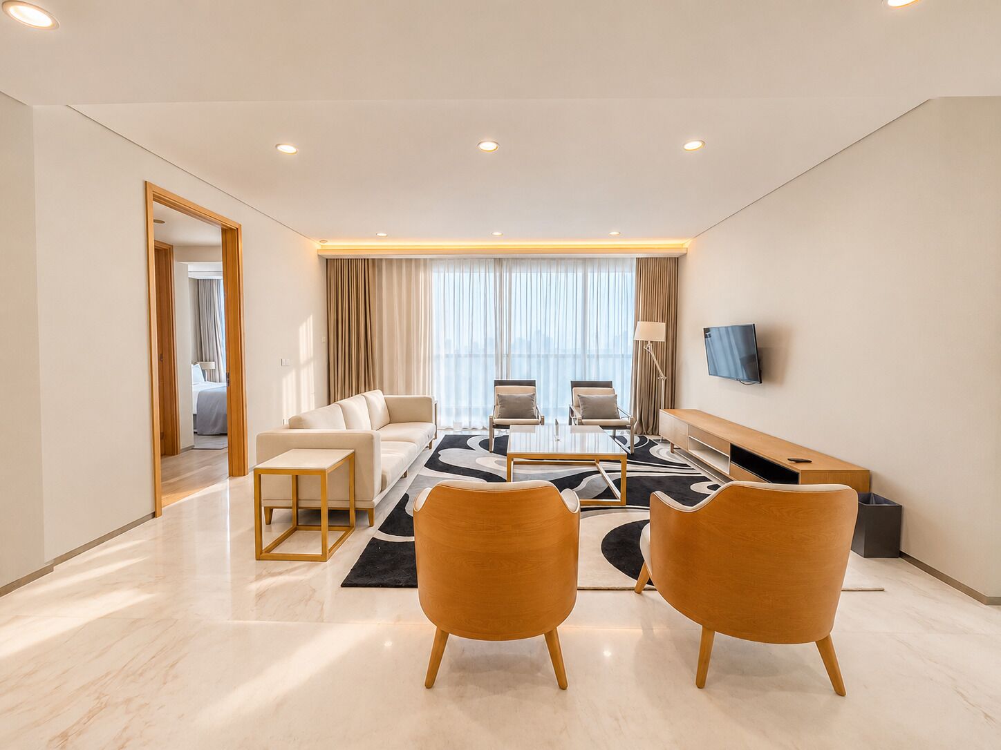 A modern hotel room with a sofa, two armchairs, a coffee table, a TV, and a view of the city.