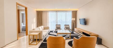 A modern hotel room with a sofa, two armchairs, a coffee table, a TV, and a view of the city.