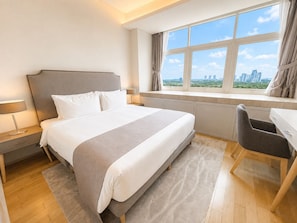 A hotel room with a bed, a desk, a chair, a window with a city view, and a nightstand with a lamp. - UHF HOTELS & RESORTS PINETREE (Iskandar Puteri)