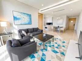 Una sala de estar moderna con un sofá, una mesa de centro y un televisor. Se observa una cocina al fondo con una mesa de comedor y sillas.