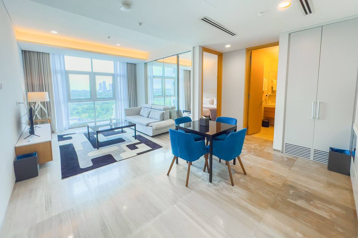 A modern hotel room with a large window, a sofa, a dining table with blue chairs, and a wardrobe.