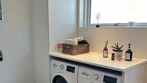 Ett kompakt tvättrum med två tvättmaskiner som har frontmatning, ett litet fönster ovanför och en hylla med toalettartiklar.