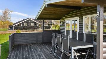 Un espacio de estar al aire libre con techo, que incluye una mesa, sillas, una terraza de madera y una vista de una casa al fondo.