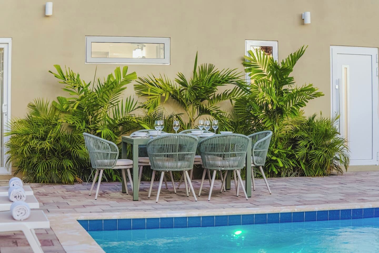 An outdoor dining area with a pool, surrounded by lush greenery and a beige wall.