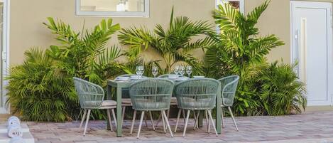 An outdoor dining area with a pool, surrounded by lush greenery and a beige wall.