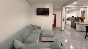 A modern living room with a sectional sofa, a flat-screen TV mounted on the wall, and a kitchen area in the background.