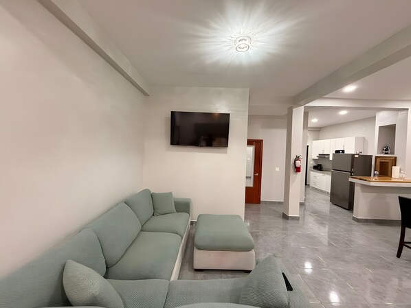 A modern living room with a sectional sofa, a flat-screen TV mounted on the wall, and a kitchen area in the background.