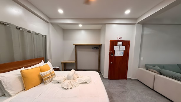 A hotel room with a bed, a sofa, a wooden table, a door with an exit sign, and a ceiling fan.