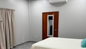 Ein Schlafzimmer mit einem Bett, einem Deckenventilator, einer Klimaanlage, einer Holztür und einem Fenster mit Vorhängen.