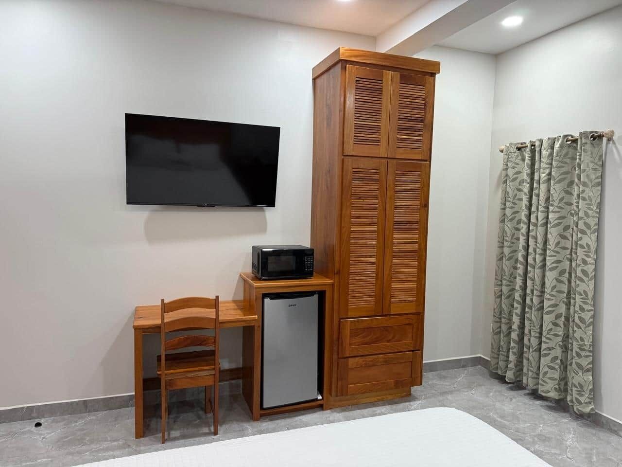 A room with a TV mounted on the wall, a wooden cabinet, a small refrigerator, a wooden chair, and a patterned curtain.