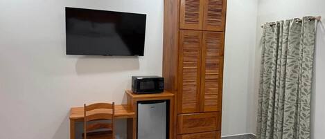 A room with a TV mounted on the wall, a wooden cabinet, a small refrigerator, a wooden chair, and a patterned curtain.