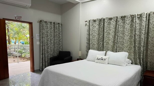 A hotel room with a bed, a chair, a nightstand, and a door leading to an outdoor area. - MeMe’s Place – Toucan (Placencia)
