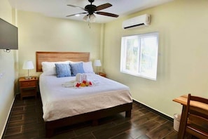 A hotel room with a bed, a ceiling fan, an air conditioning unit, a window with blinds, a TV, and a wooden chair.