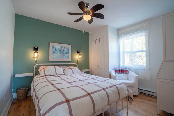 A bedroom with a bed, a ceiling fan, two wall-mounted lamps, a window, and a white chair.