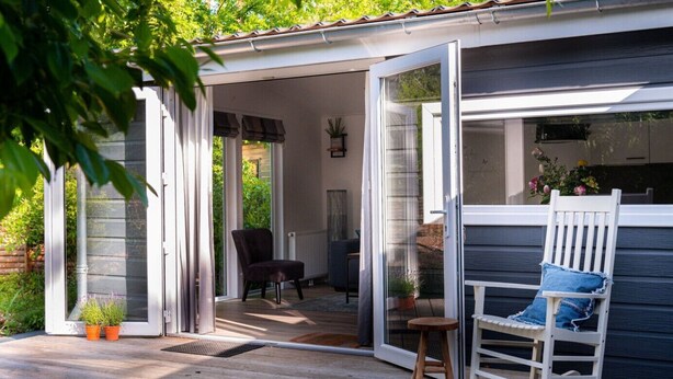 Une terrasse en bois avec une chaise berçante et une table, une porte-fenêtre coulissante donnant sur un patio, et un petit jardin agrémenté de plantes en pot.