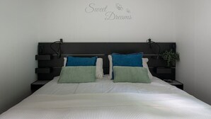 Un lit bien fait, recouvert d’une couette blanche et agrémenté de deux oreillers, un bleu et un vert, dans une chambre moderne avec une tête de lit sombre et un panneau mural sur lequel on peut lire : « Doux rêves ».