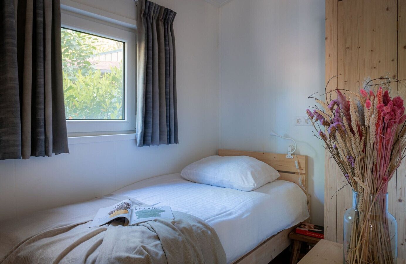 En enkelt seng med trægavl, et vindue med gardiner og en vase med tørrede blomster på et natbord i træ.
