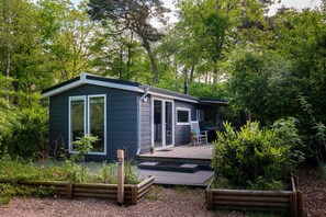 Une petite cabane aux couleurs sombres, dotée d’une terrasse et d’un espace extérieur aménagé et abrité, le tout entouré d’une végétation luxuriante.