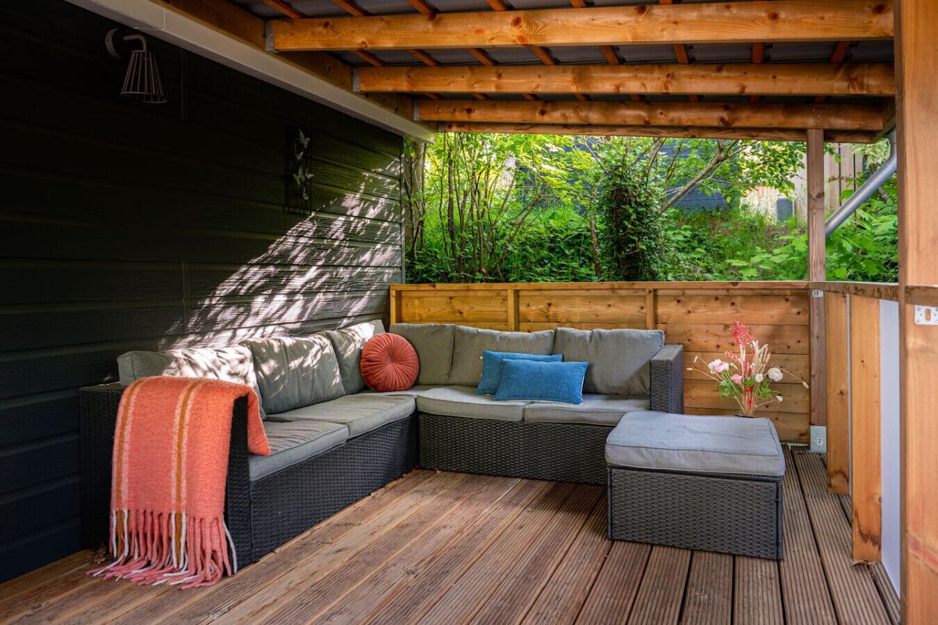 A covered outdoor seating area with a wooden deck, a cushioned bench, and a small table with a vase of flowers.