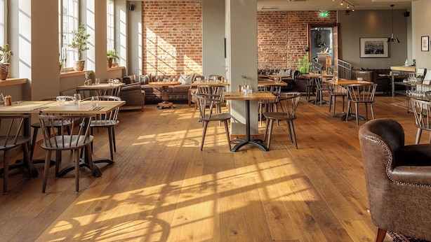 Een ruime eetruimte met houten tafels en stoelen, grote ramen voor natuurlijk licht, en een bakstenen muur op de achtergrond.