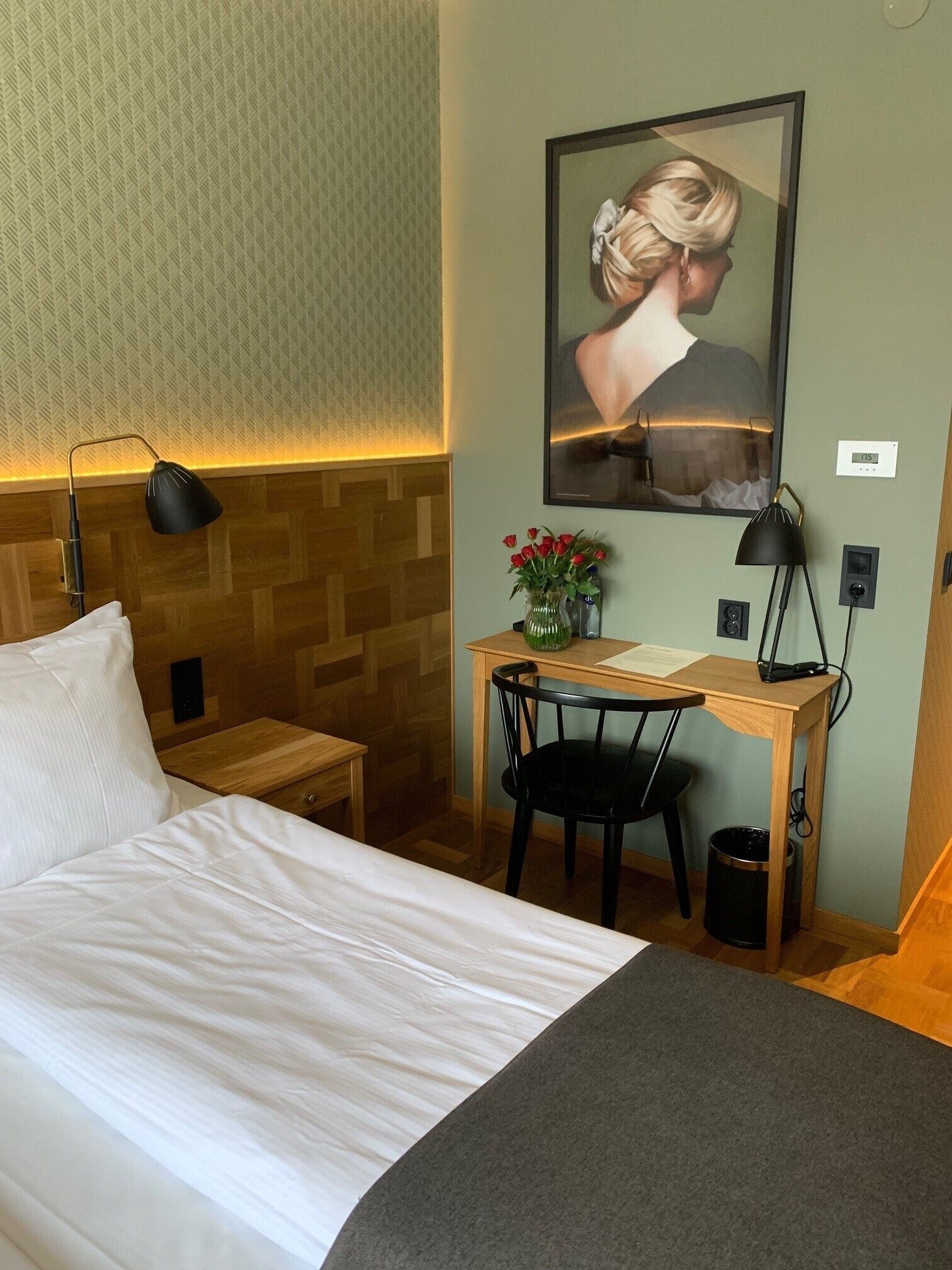 Una habitación de hotel con cama, escritorio, silla, lámpara, un jarrón con flores y un cuadro en la pared.