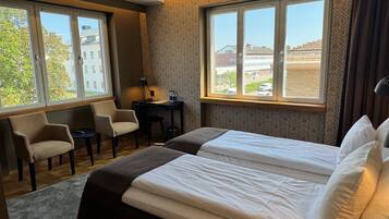 A hotel room with two beds, a desk with a chair, a lamp, and a view of buildings through large windows.
