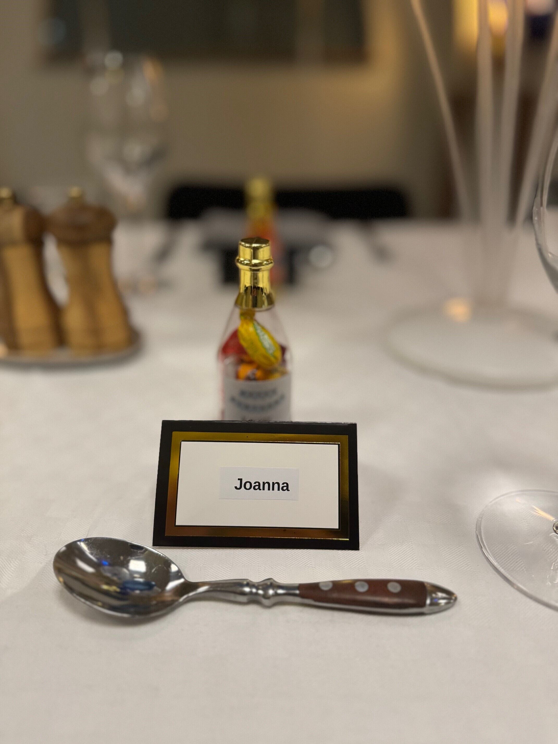 A table setting with a name card labeled "Joanna," a spoon, and a bottle of sauce.