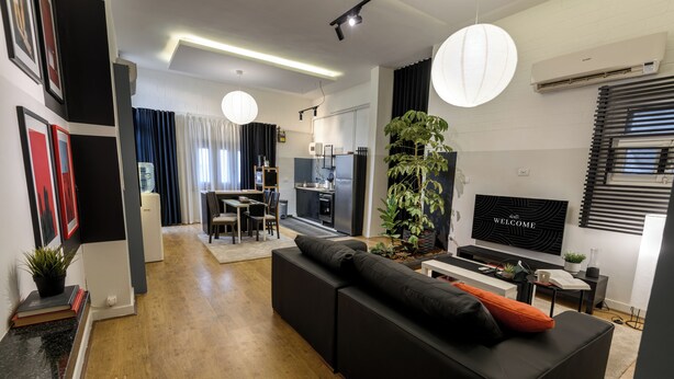 Ein modernes Wohnzimmer mit einer schwarzen Couch, einem Essbereich mit Tisch und Stühlen, einem Fernseher und einer großen Pflanze.