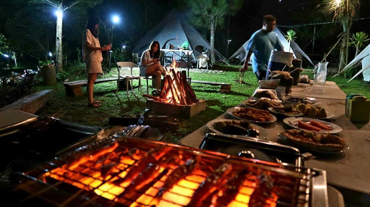 Un área de comedor al aire libre con una parrilla, mesas y sillas, rodeada de tiendas de campaña y árboles.