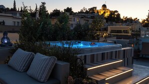 En tagterrasse med jacuzzi, udendørs møbler og udsigt over bybilledet i skumringen.