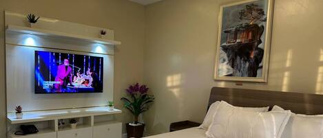 Habitación de hotel con una cama grande, un televisor de pantalla plana montado en la pared, una mesita de noche con una planta y un cuadro sobre la cama.
