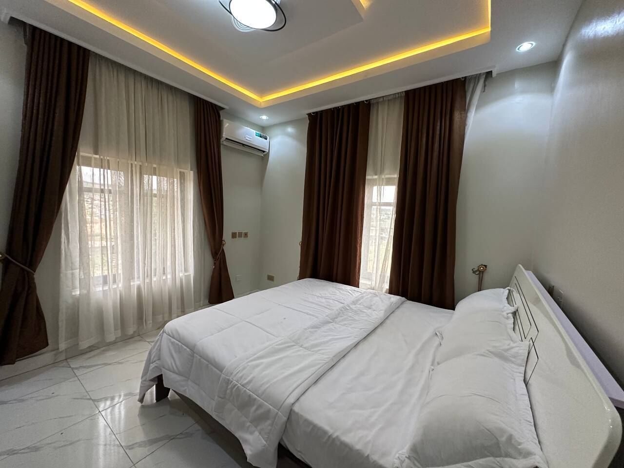 A bedroom with a bed, curtains, an air conditioning unit, and recessed lighting.